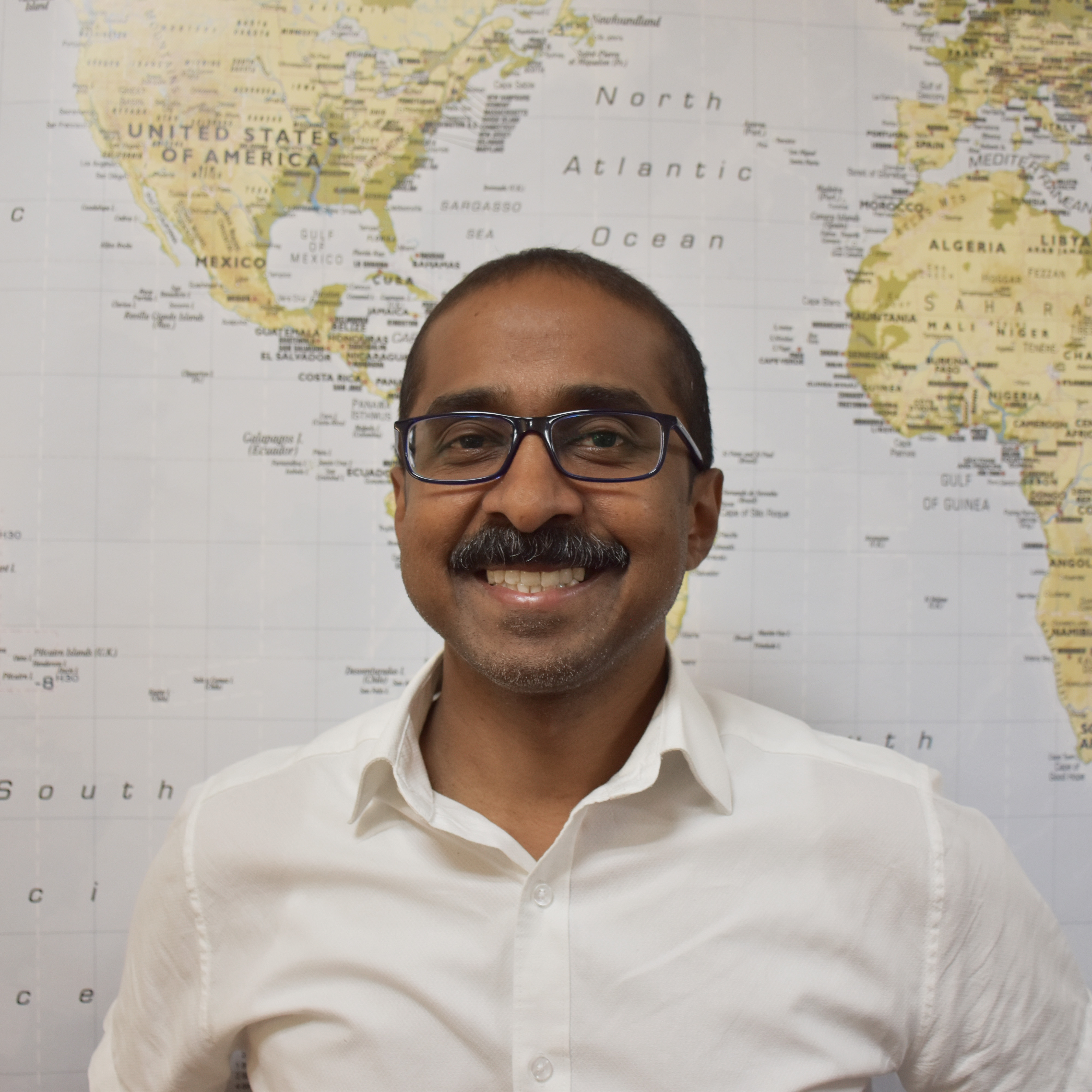 Jay Arasan, Associate Director, smiling in front a world map